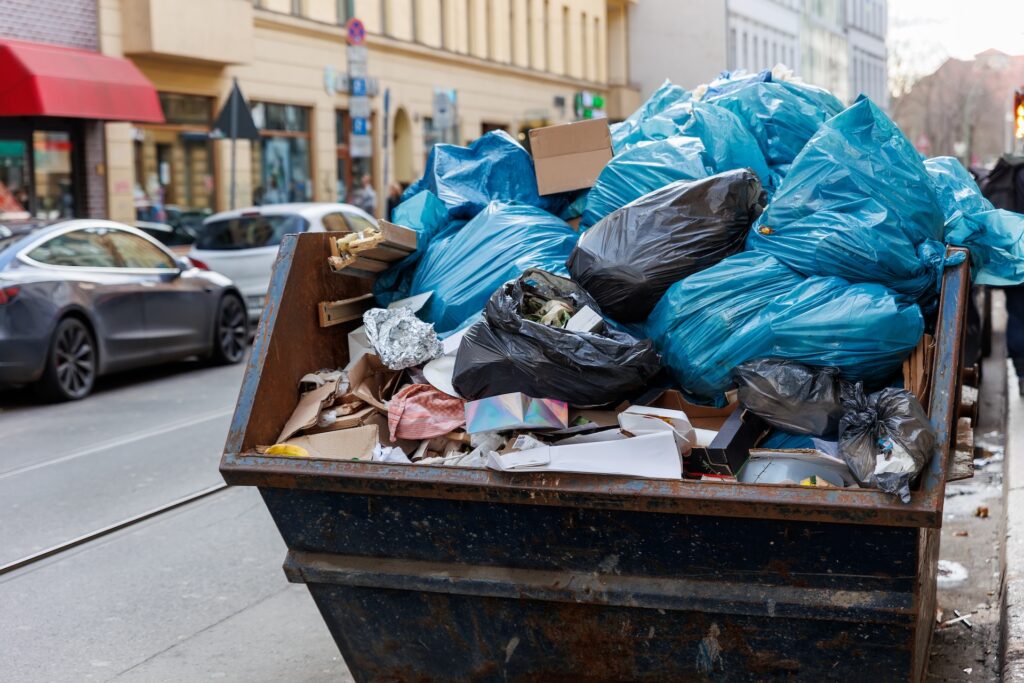 Big metal overloaded dumpster skip container full of plastic trash litter bags near residential building at Berlin city Mitte downtown or suburban area. Non-recyclable sorting garbage disposal.
