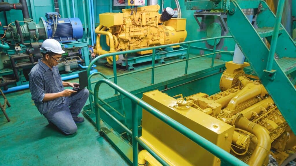 Maintenance engineer using a laptop to inspect an industrial generator system for quality and performance in a plant setting.