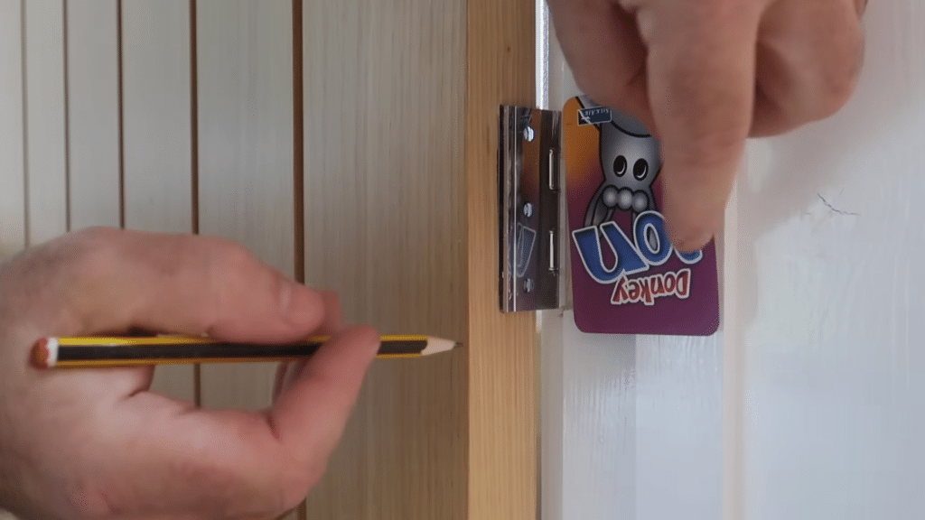 A person uses a yellow pencil to mark a wooden door jamb next to a metal door hinge secured by a purple card