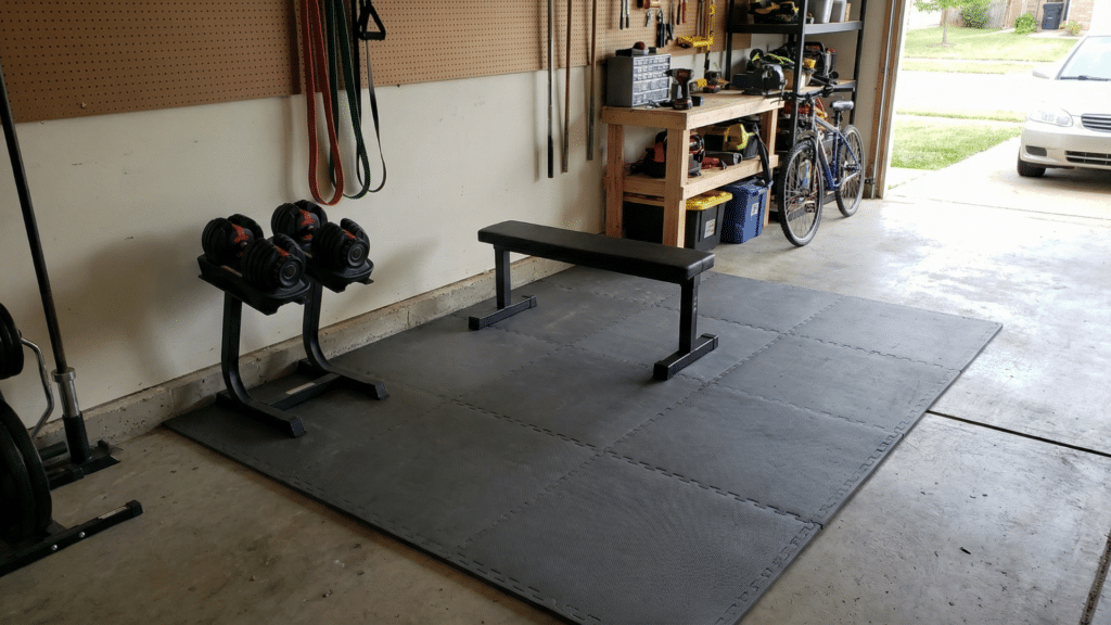 Affordable half garage gym setup with adjustable dumbbells, flat bench, and rubber tile flooring