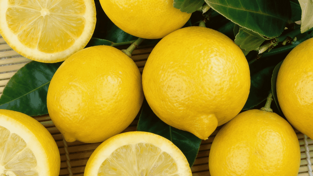 Bright yellow whole and halved lisbon lemons with dark green leaves rest on a textured, light brown surface.