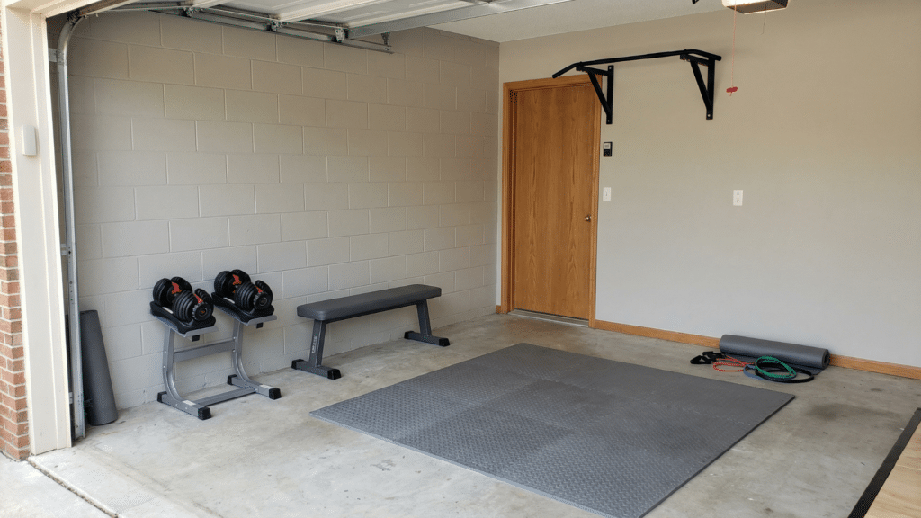 Compact half garage gym featuring adjustable dumbbells, bench, pull up bar, and open workout floor space