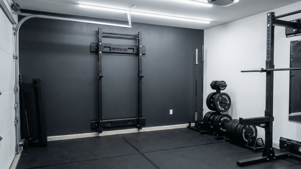 Half garage gym with wall mounted folding squat rack, barbell, bumper plates, and vertical bench storage