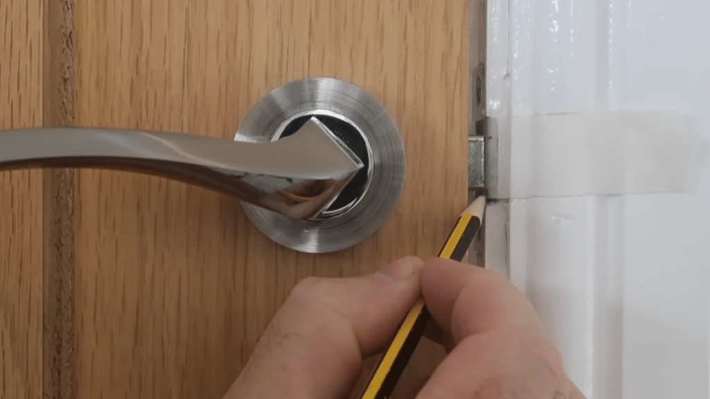 Hand holding a pencil marking the strike plate area on a white door frame adjacent to a wooden door with a chrome lever handle