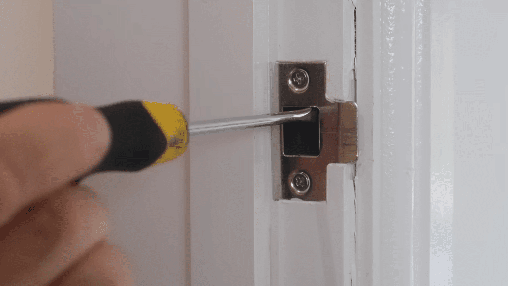 Hand using a flathead screwdriver to adjust the mechanism of a door hinge mounted on a white frame.