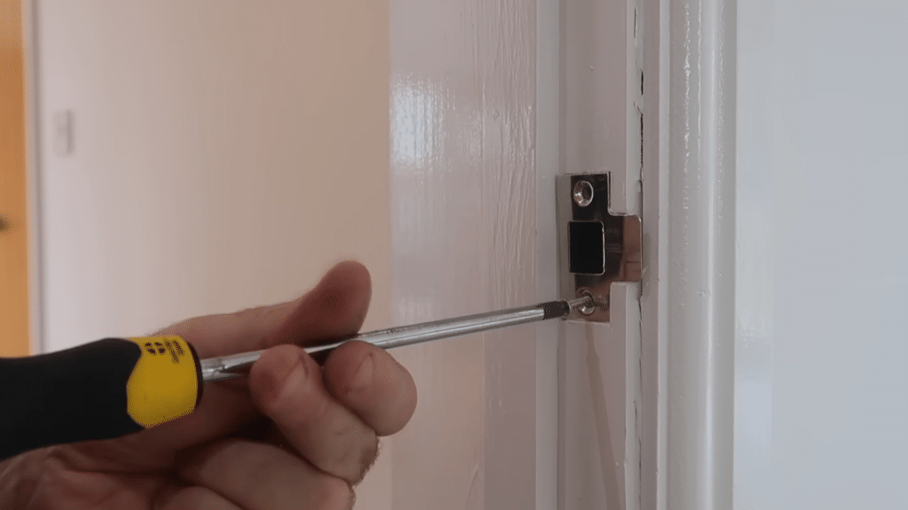 Hand using a screwdriver to install or adjust a metal door latch strike plate on a white door frame