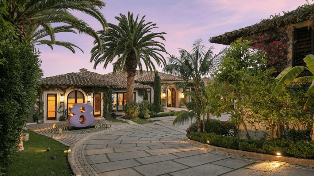 Prince Harry's home entryway featuring a large purple number eight sculpture, palm trees, and lush landscaping at twilight