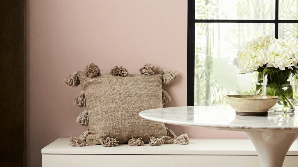 Sashay Sand SW 6051 wall color in a softly lit room with a neutral tassel pillow, white console table, marble side table.