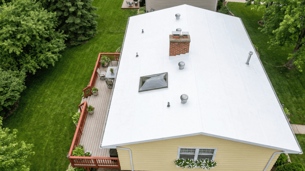 Aerial view of a house with a bright white reflective roof, a brick chimney, a skyligh , and an adjacent wooden deck surrounded by lush green lawn and tree .