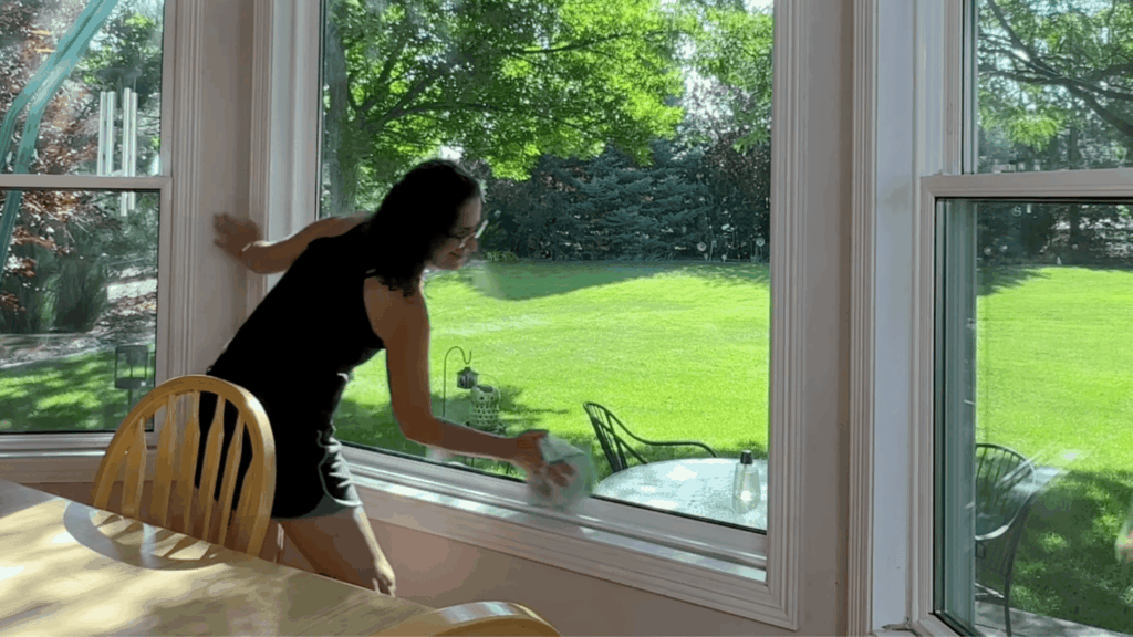 a woman cleaning large window with cloth in bright room overlooking green yard with trees and patio furniture