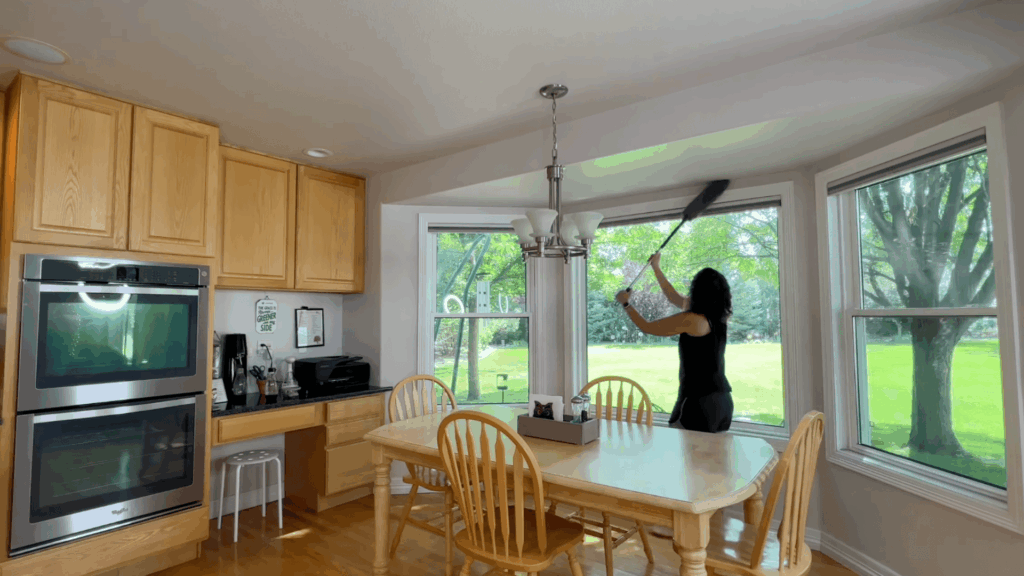 a woman cleaning large window with duster in bright kitchen dining area with wooden table chairs and green yard outside