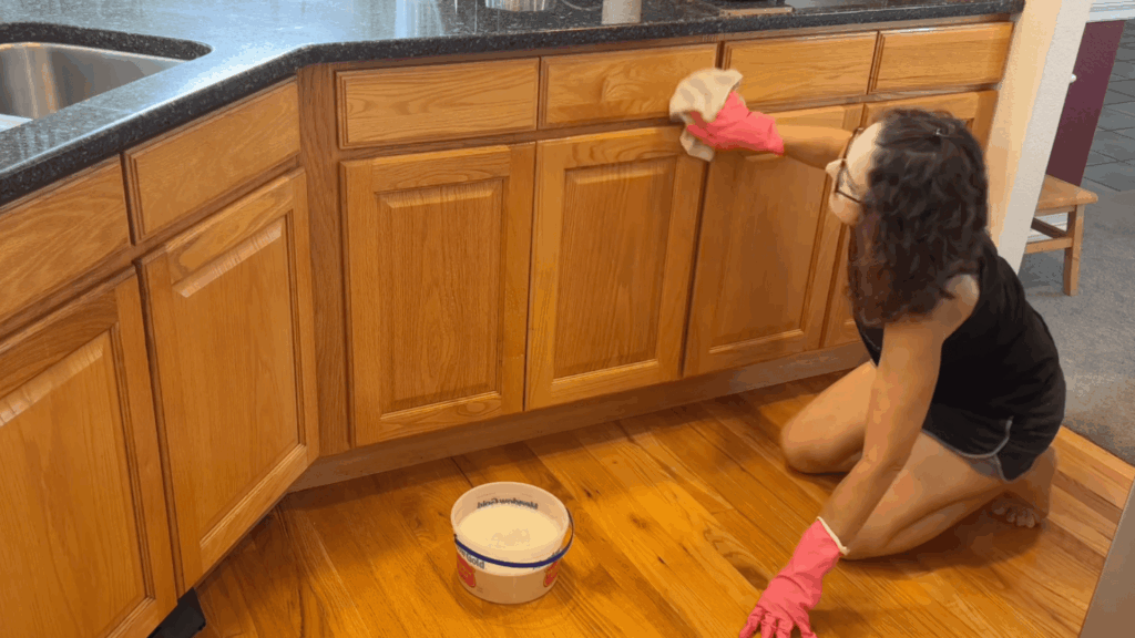 a woman kneeling on wooden floor cleaning kitchen cabinets with cloth wearing pink gloves and bucket nearby (1)