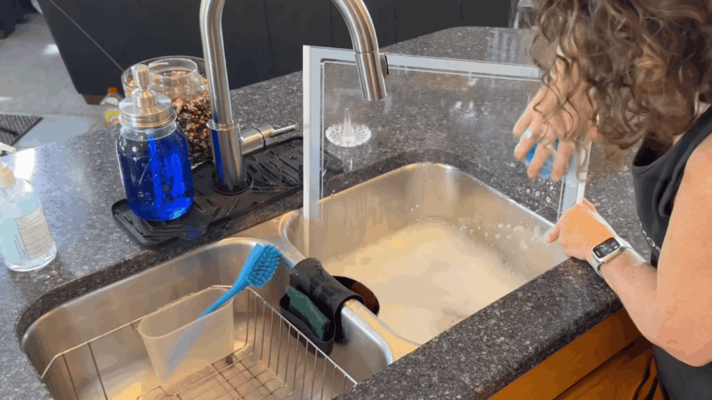 a woman rinsing glass panel in kitchen sink with running water soap bubbles and cleaning tools on countertop