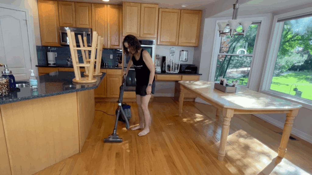 a woman vacuuming wooden floor in bright kitchen near table and windows with green yard visible outside
