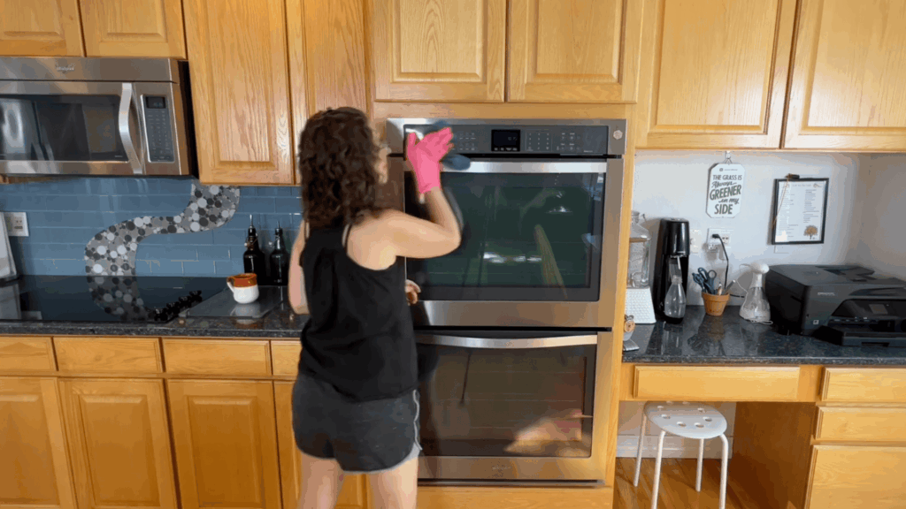 a woman wearing pink gloves cleaning oven door in kitchen with wooden cabinets countertop appliances and tiled backsplash
