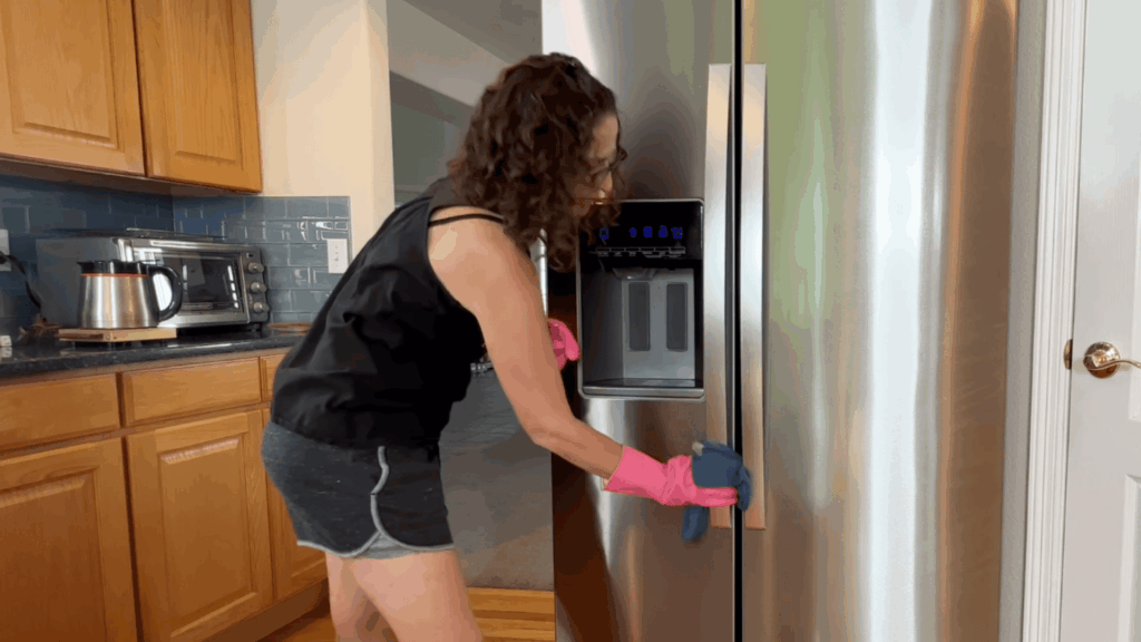 a woman wearing pink gloves cleaning stainless steel refrigerator door in kitchen with wooden cabinets and appliances