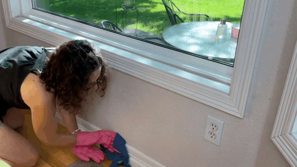 a woman wearing pink gloves wiping wall near window and floor vent in bright room with outdoor patio visible through glass