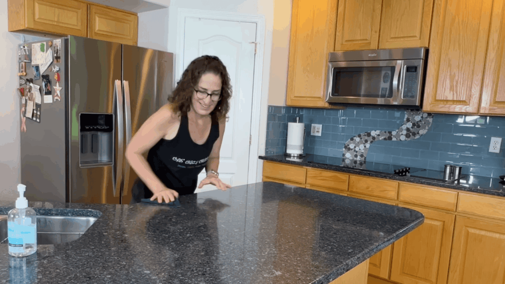 a woman wiping kitchen countertop with cloth in bright kitchen with wooden cabinets stainless appliances and sink nearby