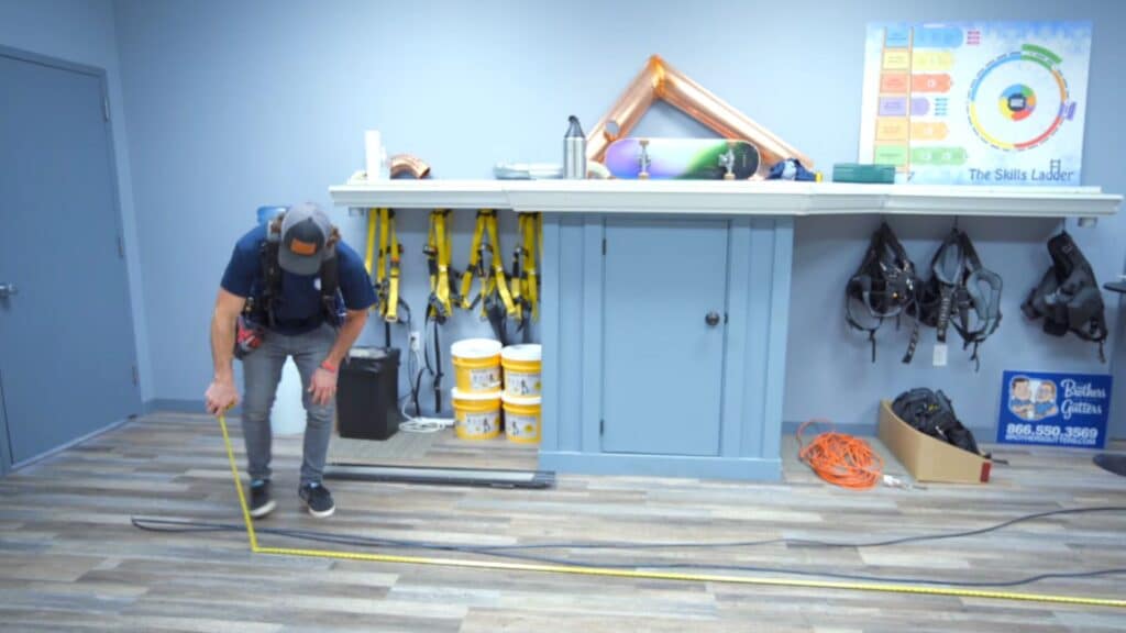 a worker kneels on floor measuring long cable with tape in workshop space with tools shelves and equipment behind