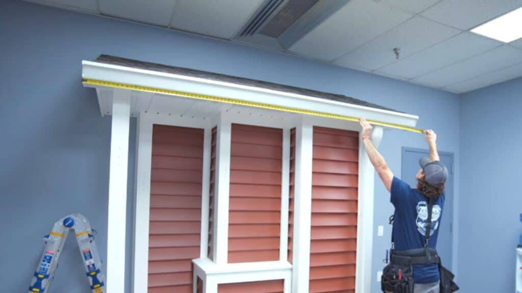 a worker measures roof edge with tape while standing beside a model house structure indoors with tools nearby