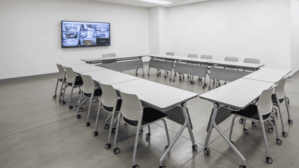 bright training room with white tables arranged in a u shape rolling chairs and a wall mounted screen in a clean modern space