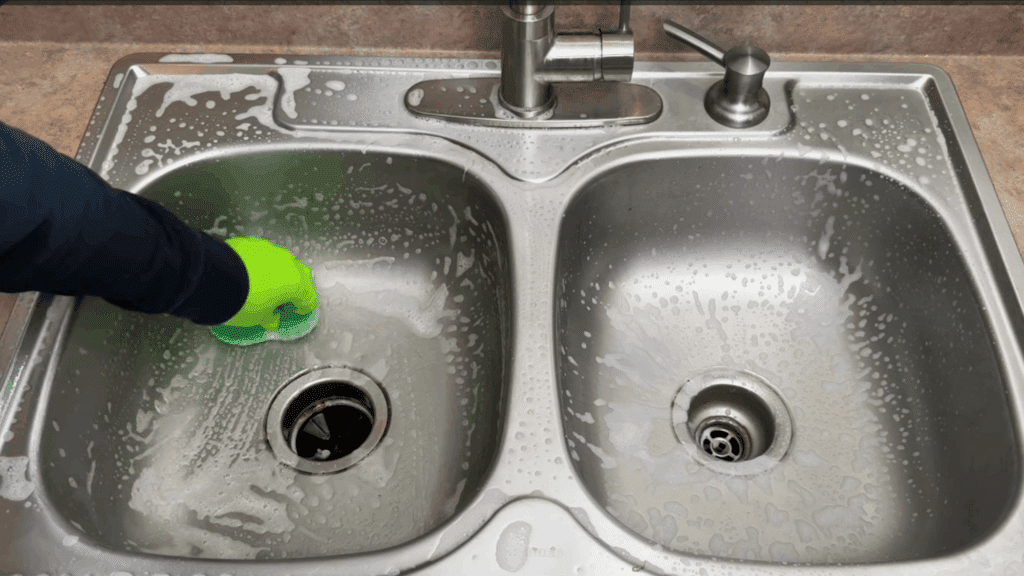 gloved hand scrubbing stainless steel sink with sponge and soap foam deep cleaning kitchen sink