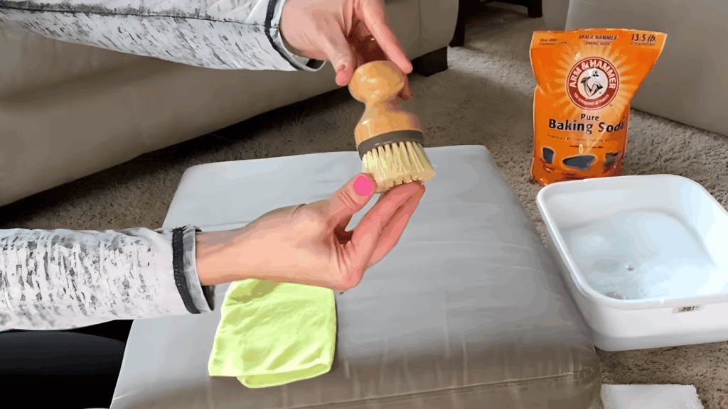 hand holding small scrub brush over cushion with cloth and cleaning mixture nearby
