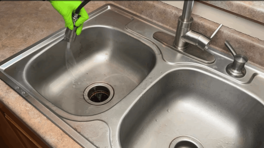 hand wearing gloves rinsing stainless steel sink with water spray cleaning kitchen surface