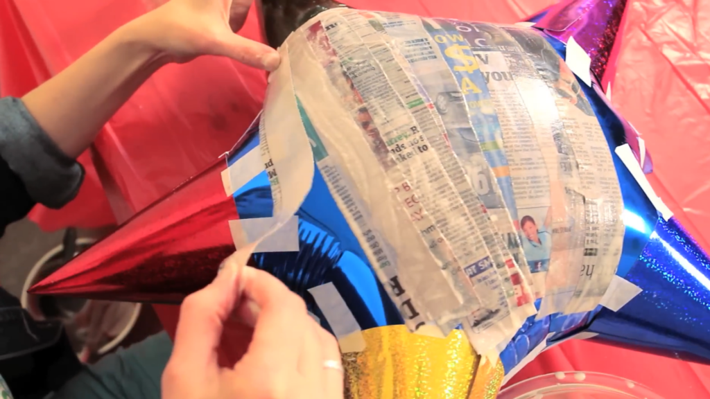 hands placing newspaper strips onto star pinata with glue layer
