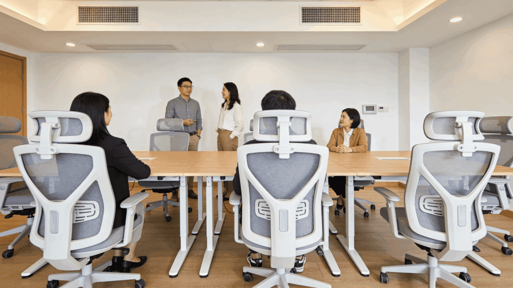 people in a meeting room sitting at a table while two colleagues stand and talk in a bright modern office setting