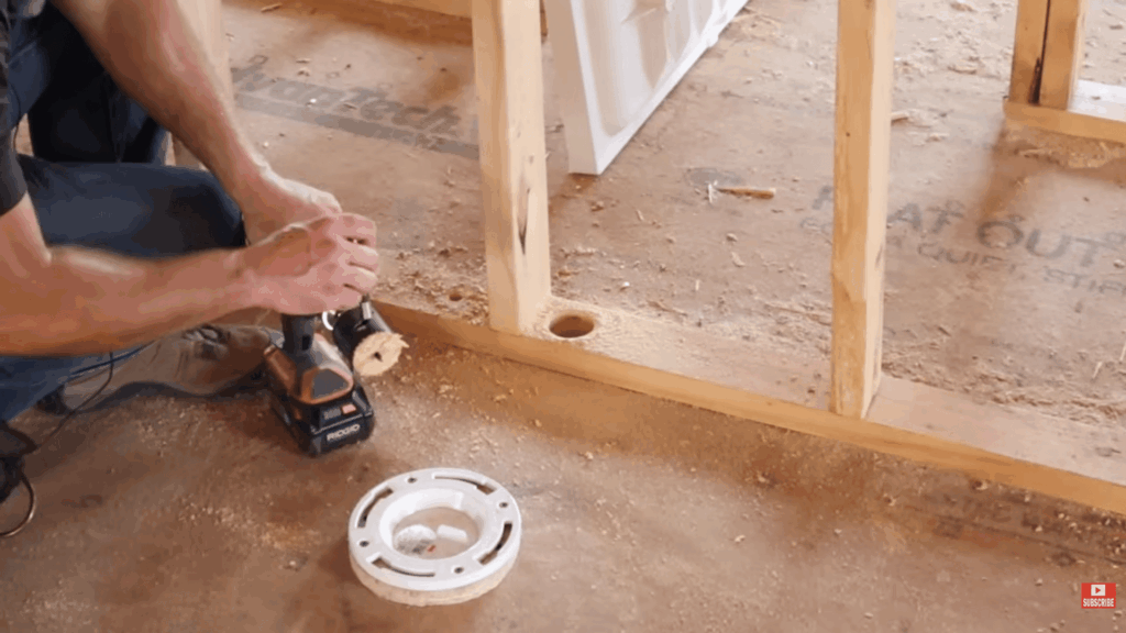 person drilling hole in wooden frame for drain pipe showing how to plumb a bathroom during installation