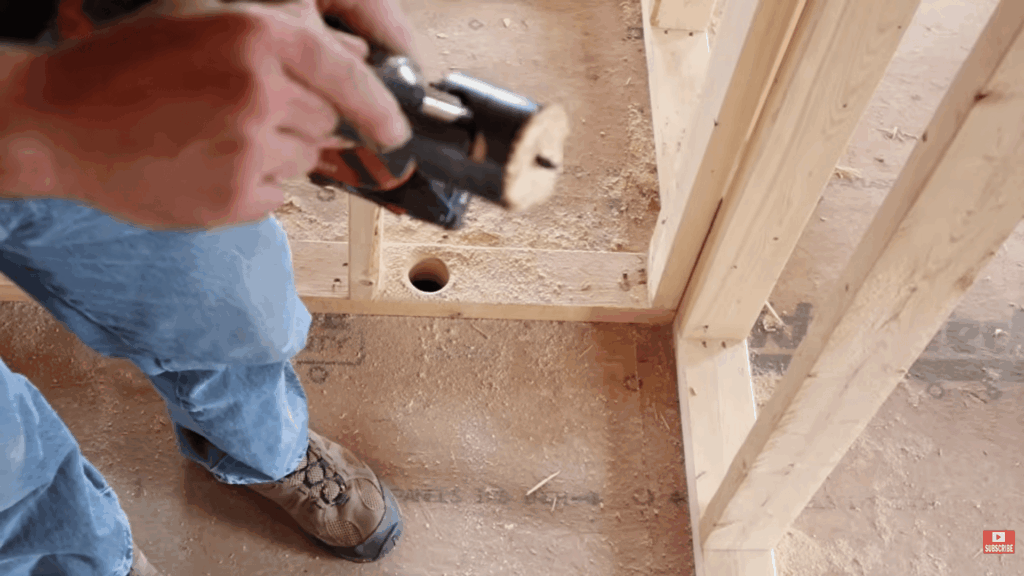 person drilling hole through wooden floor frame for pipes showing how to plumb a bathroom during installation