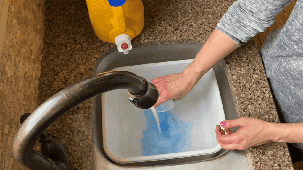 person pouring blue soapy water into white container under kitchen faucet at sink
