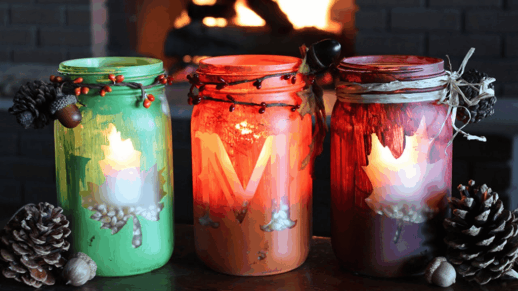 three painted mason jar candle lanterns with leaf designs glowing near fireplace for homemade gifts made easy