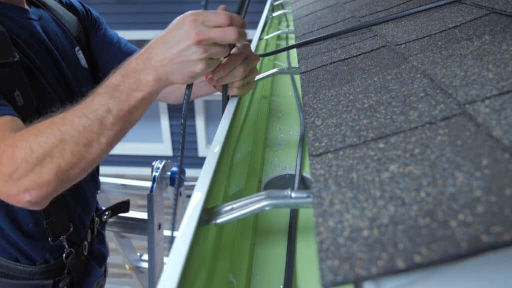 worker attaches cable along roof edge near gutter using hands while standing on ladder beside shingled surface