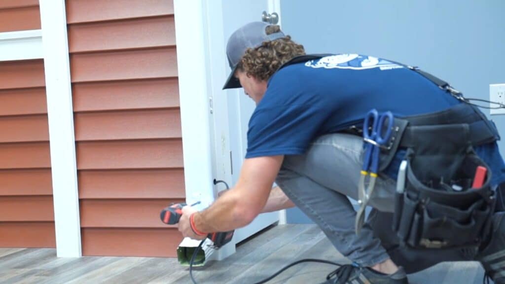 worker kneels near wall using drill to secure cable or fixture at base of structure wearing tool belt indoors