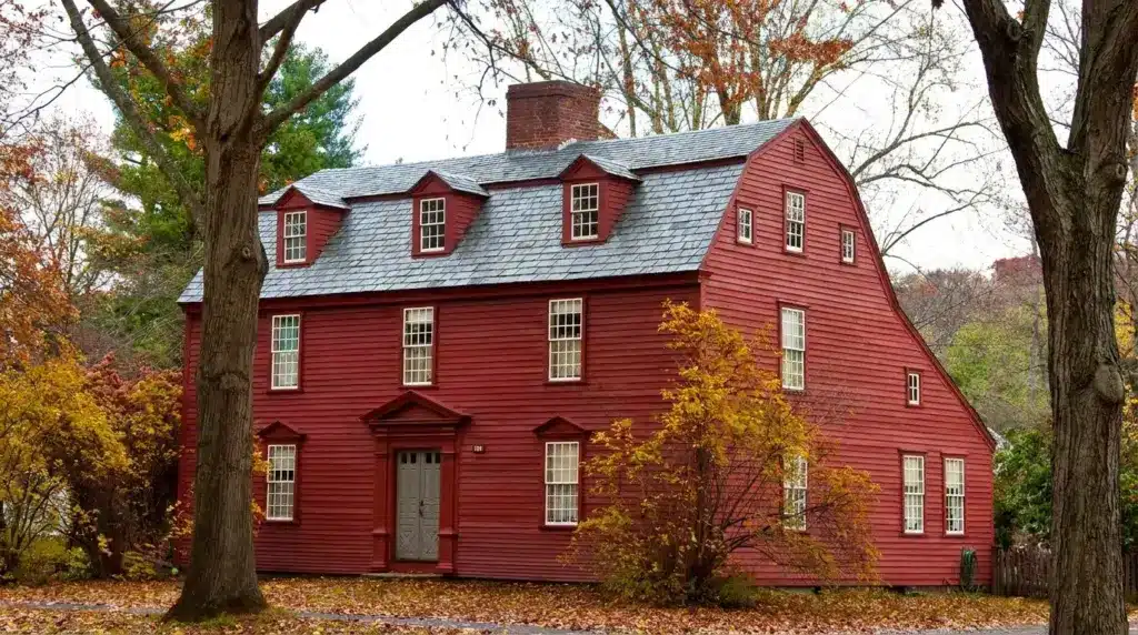 A historic red wooden saltbox house with a central chimney and small windows, nestled among autumn trees.