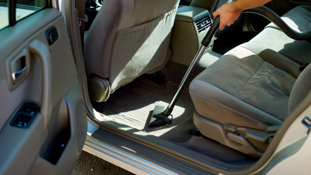 A person using a vacuum cleaner attachment to clean the light-colored fabric seats and floor mats of a car.