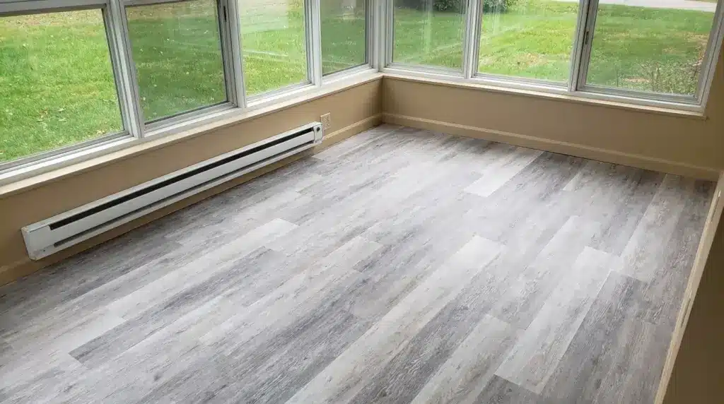 A room with large windows, featuring a light gray wood-like floating floor and a baseboard heater along the wall.