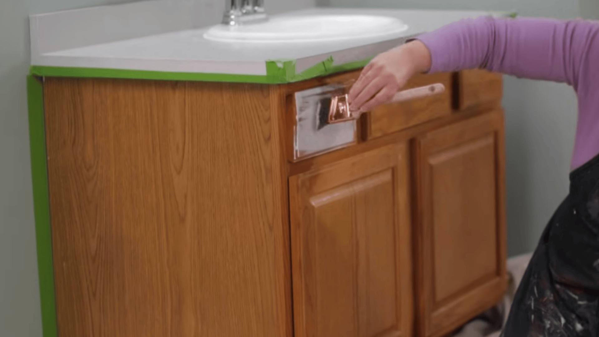 a person applies white paint to a wooden cabinet using a brush while green tape protects the counter edges