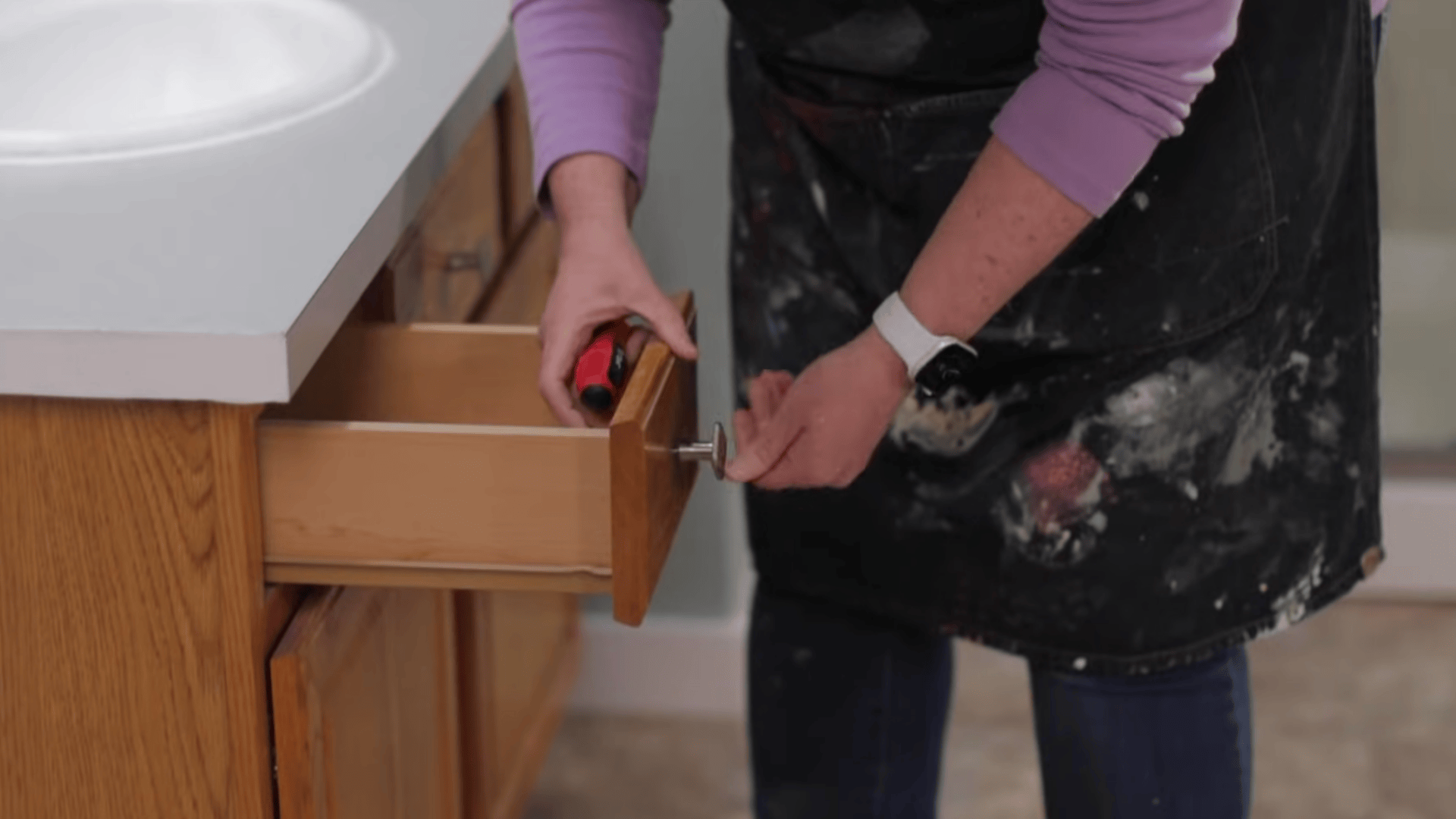 a person in stained apron opens a wooden drawer while holding a red screwdriver near a sink