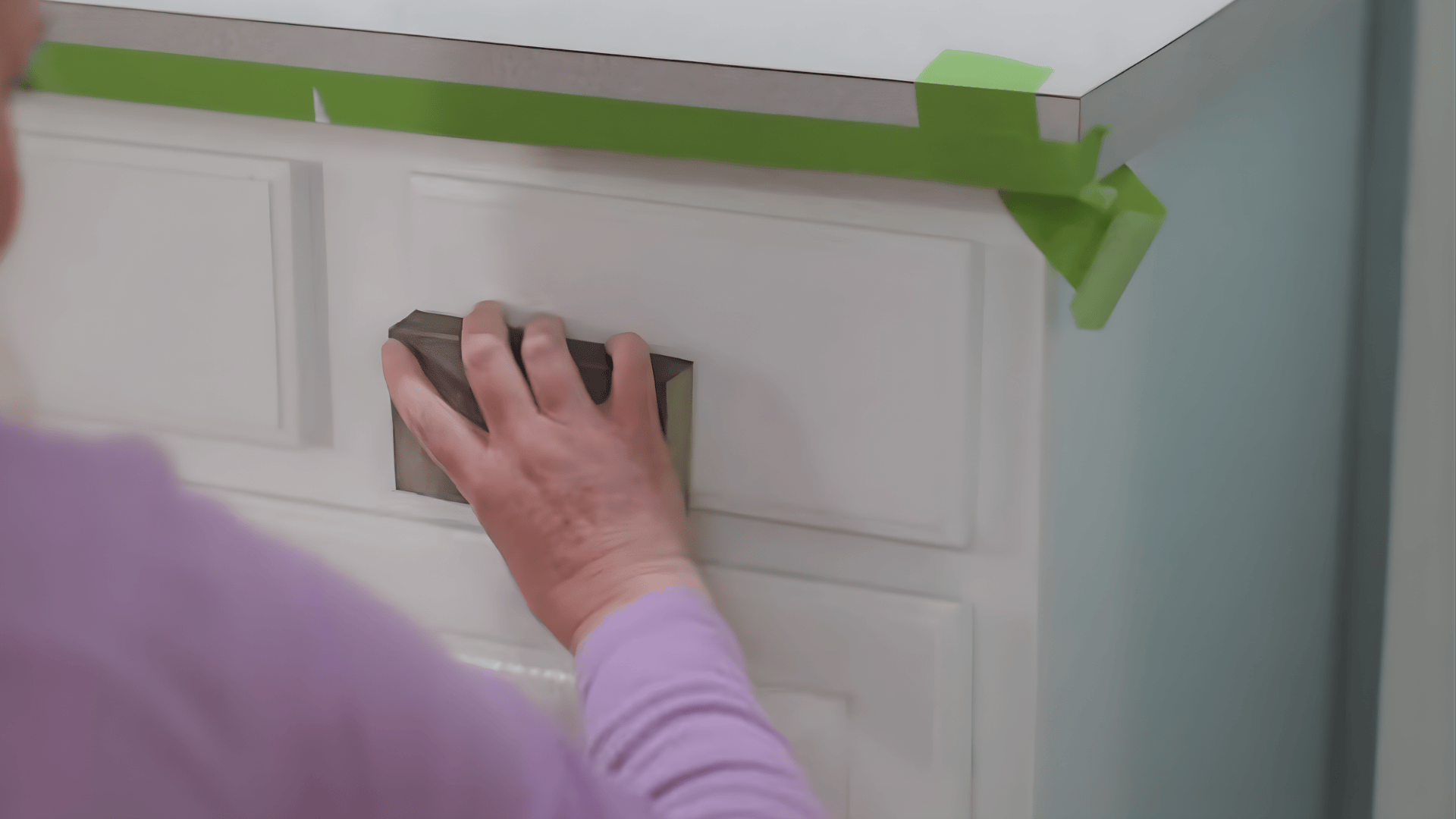a person sands a painted cabinet door with a sanding block while green tape lines the countertop edge