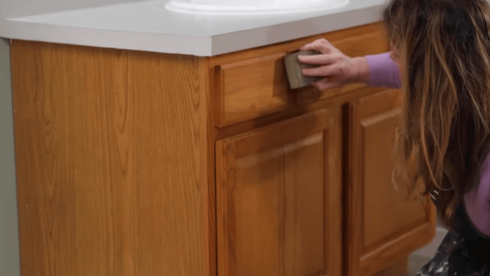 a person sands a wooden cabinet drawer front with a sanding block beneath a white countertop near a sink