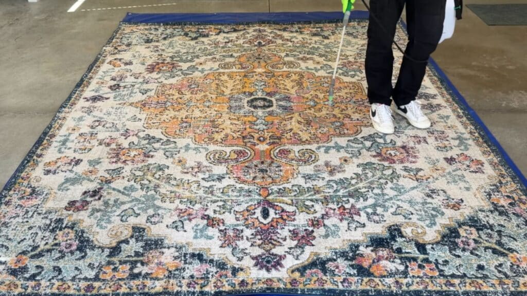 a person spraying cleaning solution evenly across a large patterned rug laid flat on a tarp indoors