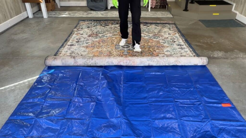 a person unrolling a patterned rug over a blue tarp on a concrete floor indoors preparing for cleaning process