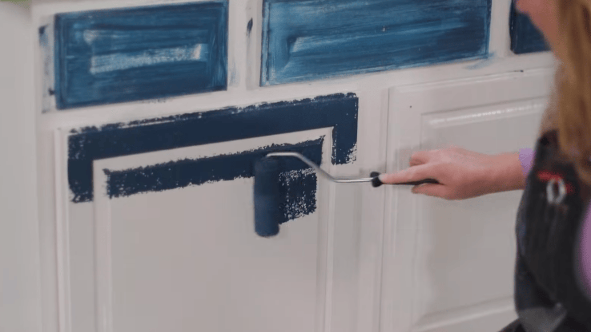 a person uses a small paint roller to apply dark blue paint onto white cabinet doors with recessed panels