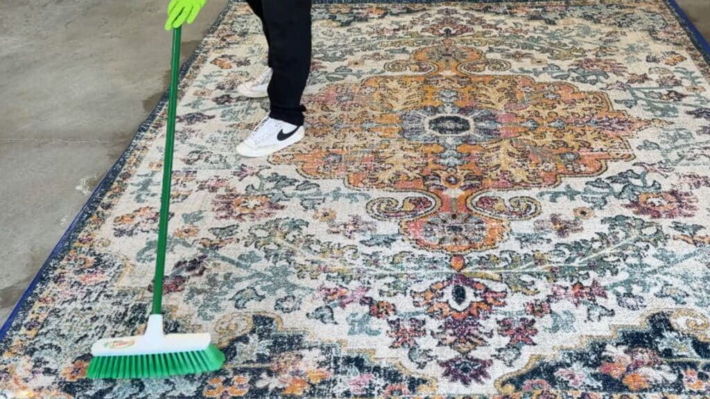 a person using a push broom to scrub and spread cleaning solution across the surface of a large rug