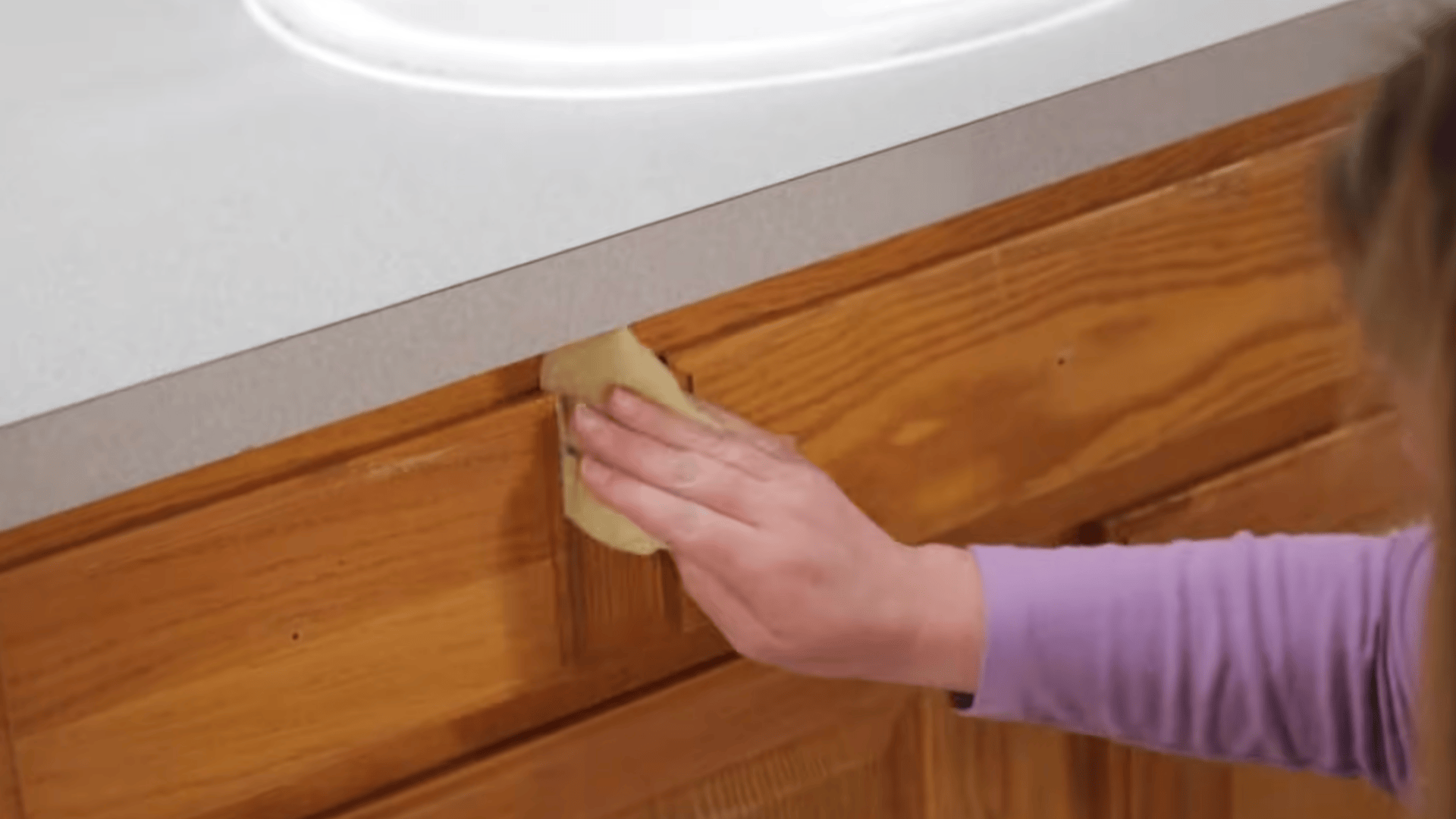a person wipes a wooden cabinet with sandpaper beneath a bathroom sink counter smoothing the surface before finishing