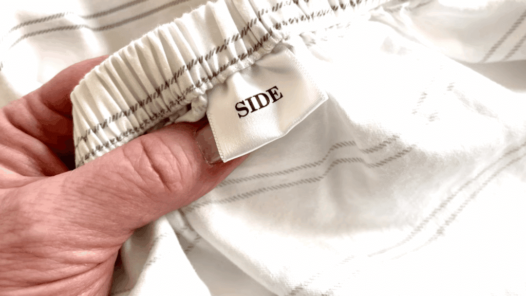 close up of hand holding fitted sheet edge with elastic band and tag labeled side on soft white striped fabric