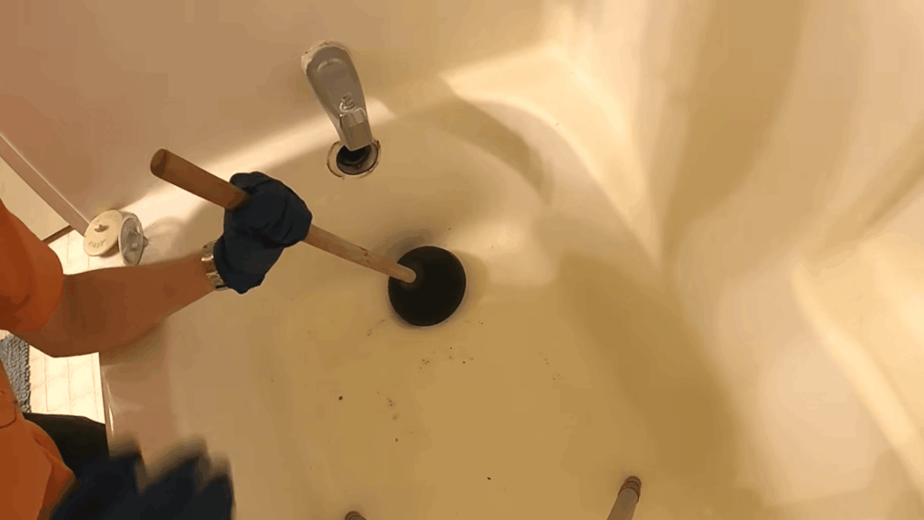 gloved hand using plunger in bathtub drain showing how to unclog a bathtub drain with simple tool method